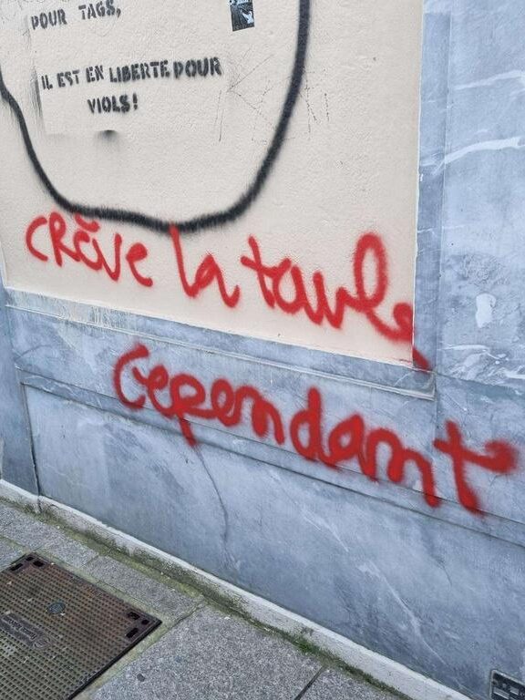 tags sur un mur, on distingue la fin d'un message "il est en liberté pour viols" et dessous, un autre tag ajouté "crève la taule cependant"