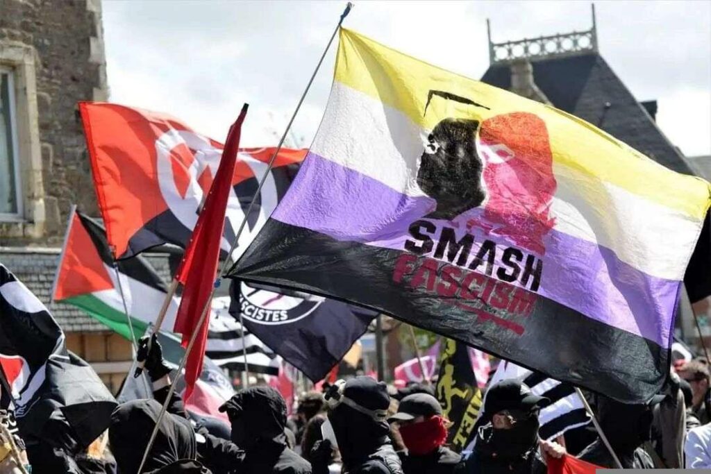 Photo d'une manif antifascite, le bloc vétu de noir. Un drapeau non-binaire flotte au vent. Peint par dessus, deux visage cagoulé en rouge et noir avec l'inscription smash fascism